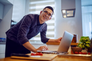 Smiling caucasian chef in uniform and with eyeglasses using looking at recipe on laptop while standing in kitchen.